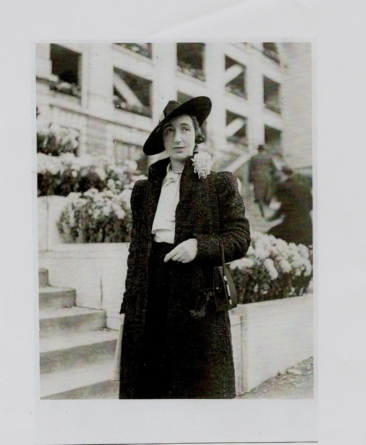 Odette en manteau noir et chapeau devant des massifs de fleurs
