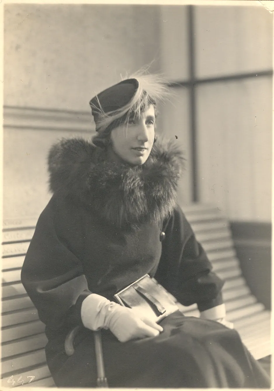 Odette en manteau à col de fourrure, assise sur un banc aux courses